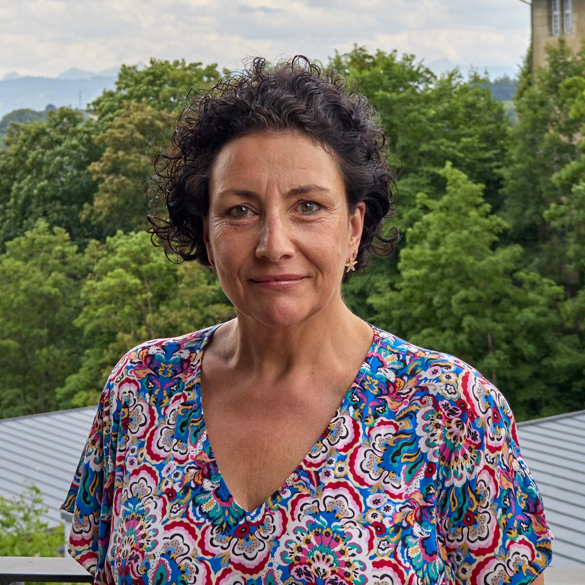 Porträtfoto von Martine Sapia. Sie trägt ein buntes Outfit und steht vor einem Gebäude..