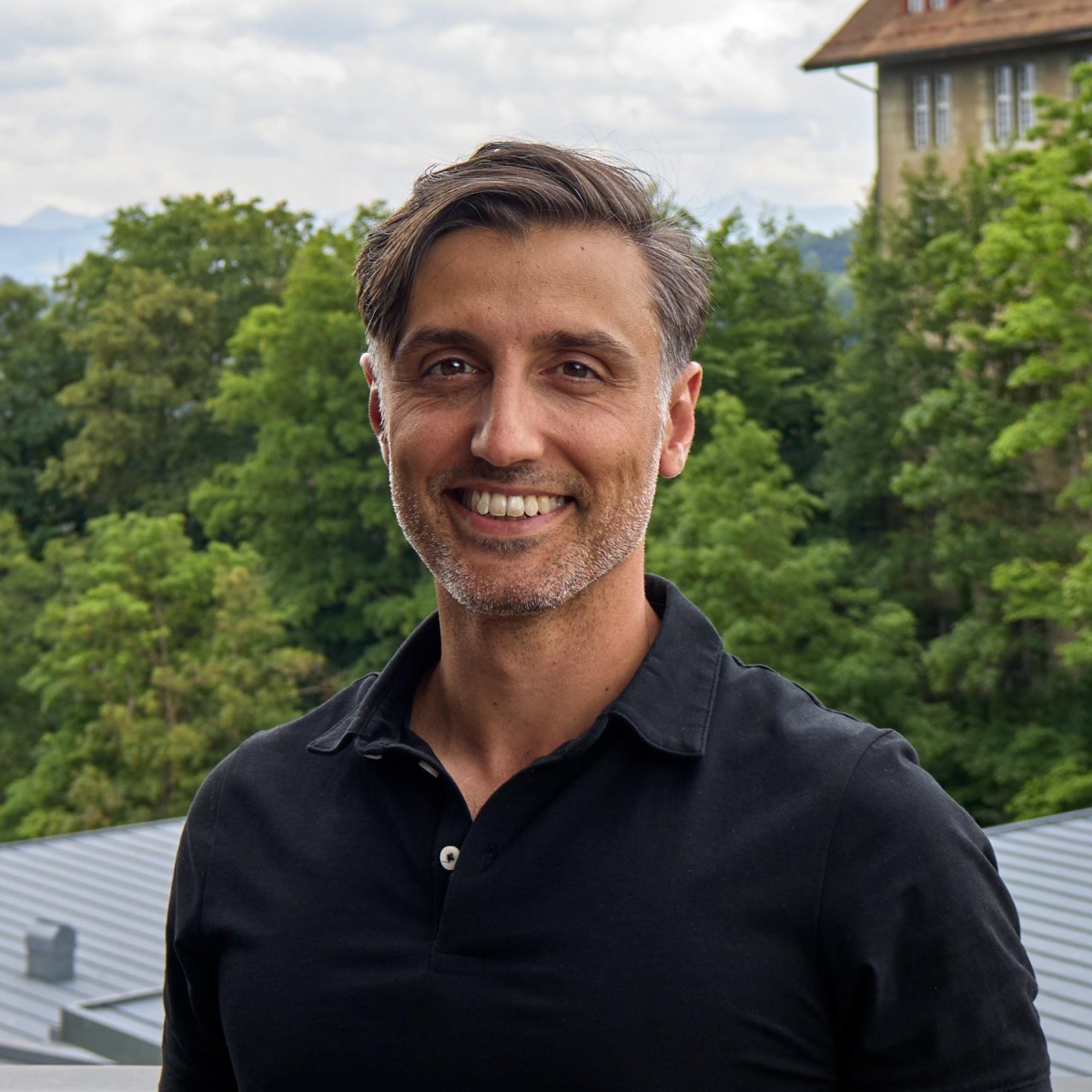 Porträtfoto von Lamb Philippe. Er lächelt, trägt ein schwarzes Hemd und steht vor einem Gebäude.