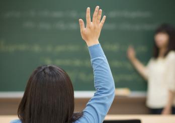 Ein Schüler in Rückenansicht hebt die Hand, um auf eine Frage seiner Lehrerin zu antworten